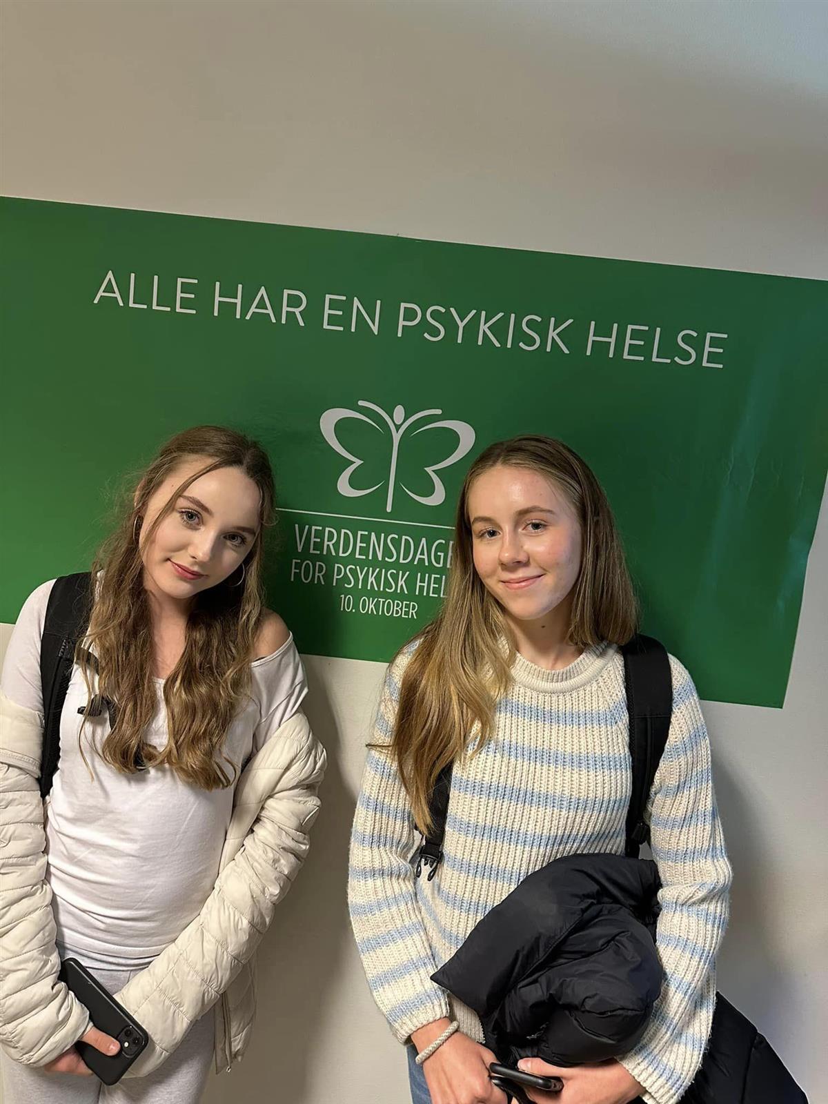 To ungdomsskolejenter med langt hår foran plakat der det står alle har en psykisk helse - Klikk for stort bilde